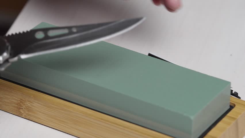 Close-up of hands sharpening a folding pocket knife blade on a green whetstone whet rock