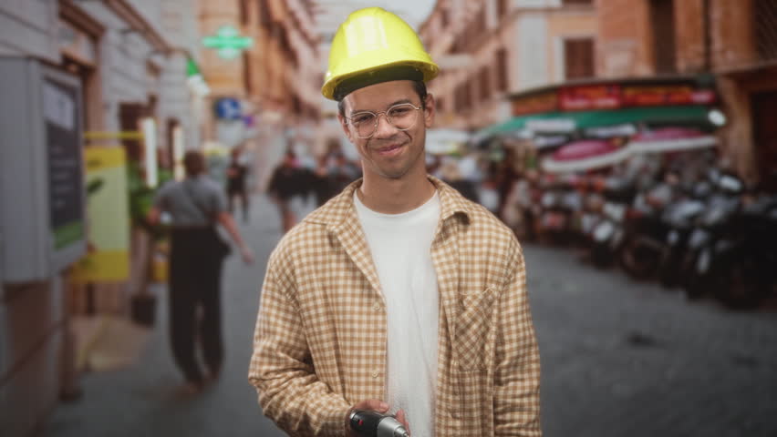 Man construction worker with yellow hardhat holding drill and pointing thumb over shoulder in street; confidence readiness.