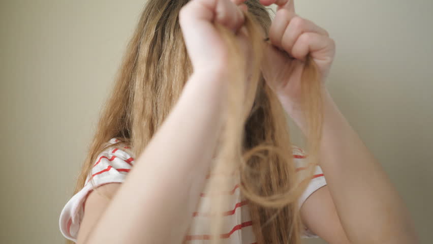 Close up of beautiful small girl weaving small pigtail out of her long blonde hair indoor. Little cute female child doing herself a hairstyle at home. Slow motion