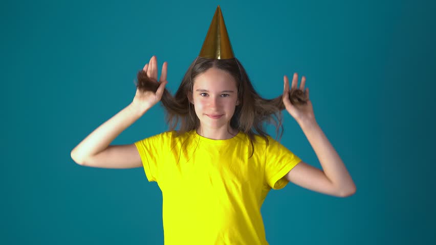 Happy cheerful girl wearing golden party cone hat and bright yellow t-shirt posing against teal blue background, holding her hair with raised hands, smiling playfully at camera. Fun birthday celebration concept