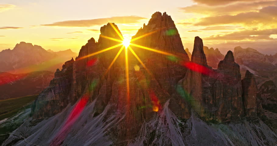 Aerial drone view of Tre Cime di Lavaredo at sunrise with dramatic sunstar between the iconic peaks. Golden morning light illuminates the rugged Dolomite landscape in northern Italy.