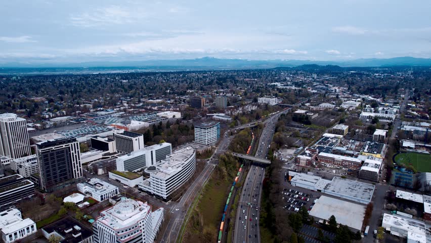 Expansive high-angle panoramic view of an urban environment. A multi-lane highway bisects the scene, surrounded by commercial buildings and industrial zones. Distant mountains meet a soft blue sky on the horizon, showcasing modern infrastructure and city planning