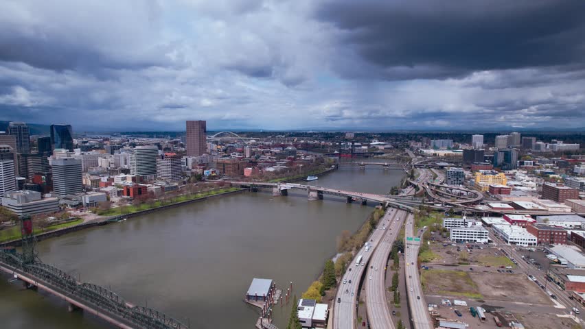 High-angle aerial view of Portland, Oregon. The Willamette River flows through the city, crossed by multiple bridges. Urban architecture, industrial zones, and busy highways are visible under a moody, overcast sky