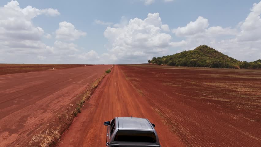 Wide view of a vehicle traveling on a long red dirt road through an arid landscape. A dust trail follows the vehicle under a blue sky with clouds near a green hill