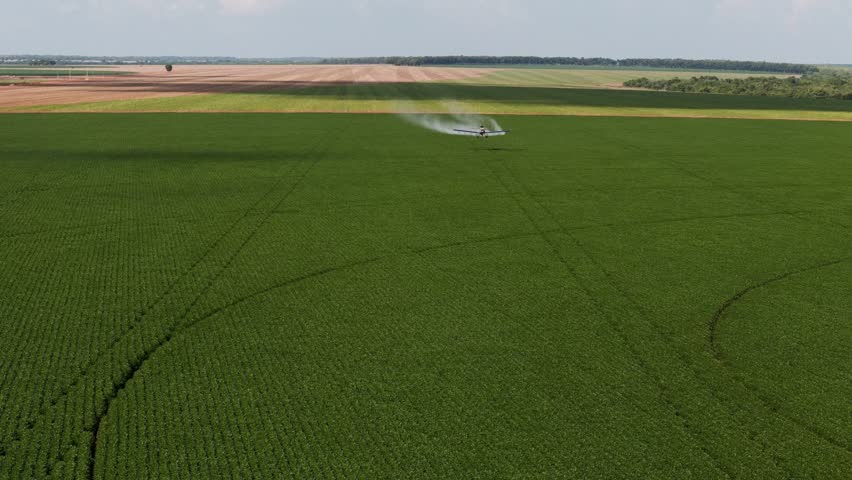 Aerial view of a crop duster airplane spraying liquid chemicals over a vast green field. The aircraft flies low, releasing a mist of fertilizer or pesticide across the plantation