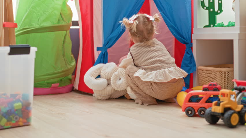 Little blonde girl hugging and lifting a large white plush octopus toy amid tents and toys in a playroom building; playful imagination.