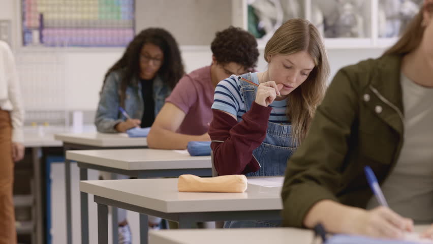 Mature teacher monitoring students as they write an exam in a classroom at secondary school. Female educator supervises multiethnic students focusing on test papers. Guys ang girls writing on exams sheets under teachers supervision at high school.