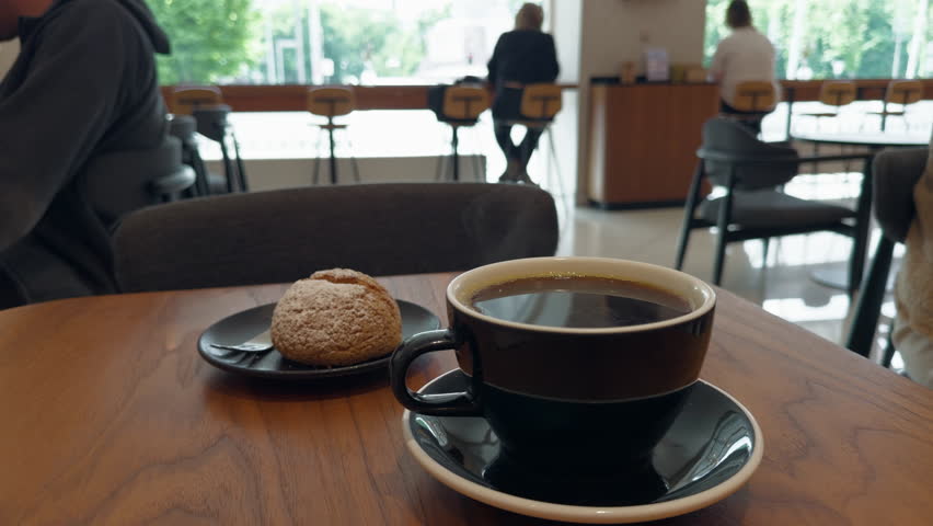 Coffee cup and pastry on table in bright modern cafe with blurred patrons in background