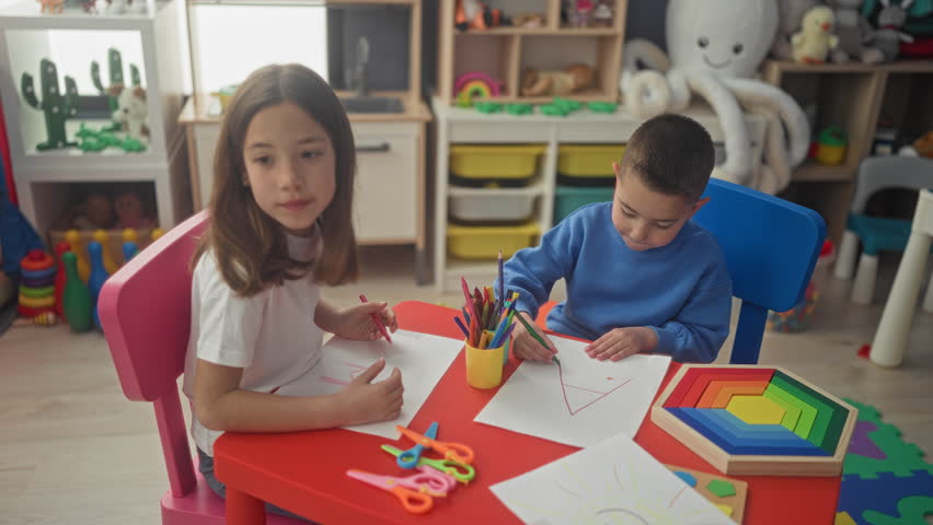 Girl and boy sitting at red kids table drawing with colored pencils on paper, scissors and wooden rainbow toy beside them inside building; creative concentration.