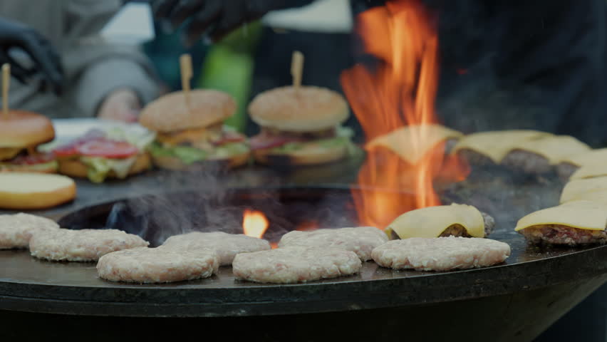 Burger patties and cheeseburgers grilling over open flames on outdoor flat top cook surface