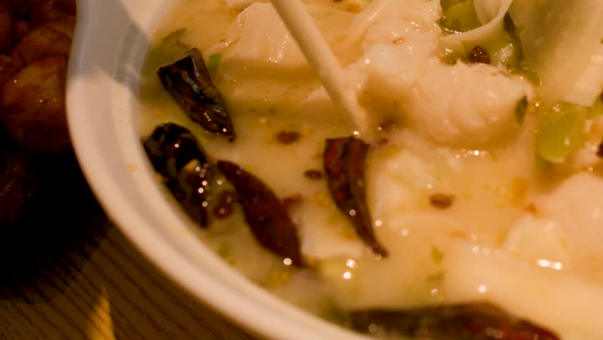 Close-up shot of chopsticks lifting fish and noodles from a spicy ceramic bowl of soup.
