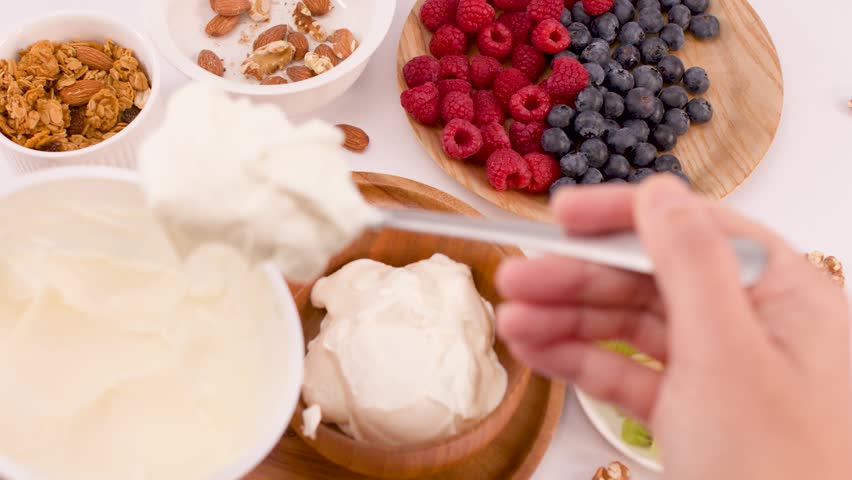 Overhead shot of hand scooping white yogurt into bowl surrounded by fresh fruit and nuts.