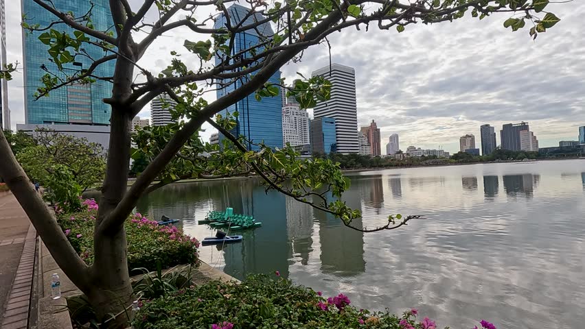 Wide panning shot of skyscrapers reflecting in a calm lake under a cloudy sky.