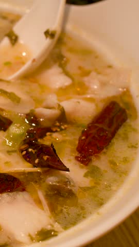 Close-up shot of chopsticks lifting white fish from spicy noodle soup in ceramic bowl.
