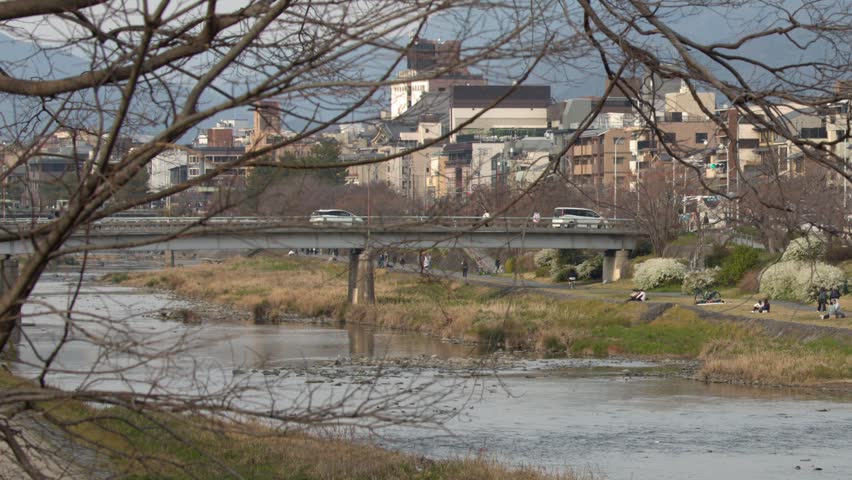 Static wide shot of a riverbank, bridge, and urban buildings under a clear winter sky.