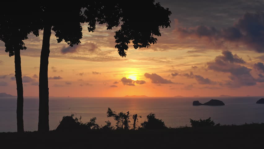 Silhouettes of two trees frame a golden hour orange sunset over tranquil ocean. Revealing small islands in the distance against a vibrant sky with scattered clouds. Tropical Thailand nature landscape
