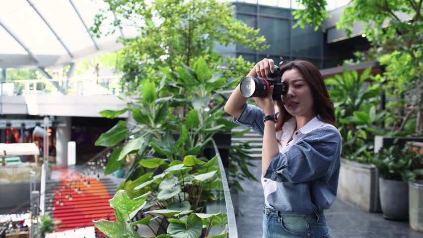 Young Asian female photographer taking pictures with a professional digital camera, reviewing the captured images on the camera screen and then looking ahead during an outdoor photo shoot.