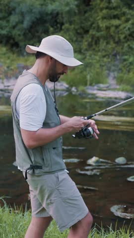 vertical Hispanic fisherman wearing a hat and vest standing on the grassy riverbank. Actively reeling in the fishing line with his rod