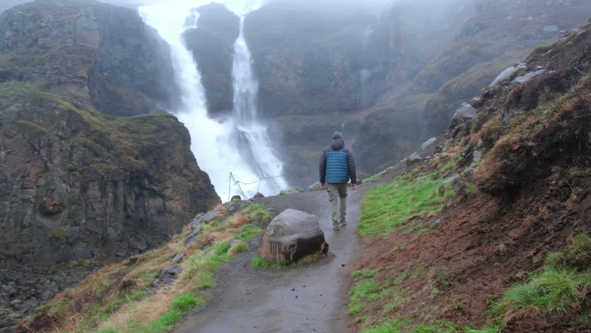 Adventurous hiker walks a rocky trail toward dramatic Rjukandafoss waterfall, mist and fog filling the canyon for a moody, powerful Icelandic landscape scene