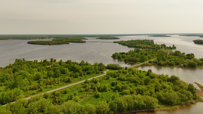 Drone side pullback revealing an island along Long Sault Parkway with the scenic roadway crossing the St. Lawrence River and additional islands in the distance.