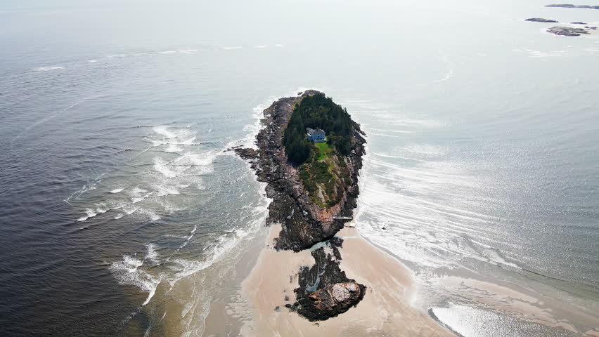 Wide aerial revealing a secluded coastal mansion on a private island near Popham Beach, Maine, with surrounding evergreen forest and a sandbar connecting the house to the protected beach coastline.