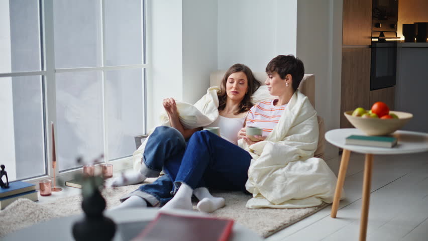 Peaceful ladies drinking coffee wrapped cozy blanket together sitting living room floor. Happy two women sipping hot beverage talking enjoying cold winter morning. Smiling lesbian couple relaxing home