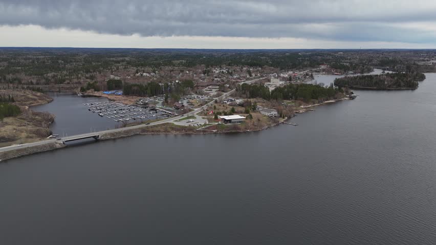 Kenora, Ontario. Dronie shot over Lake of the Woods backs away from Canadian flag, revealing marina, town of Kenora, lakeside highway, cars, boats, and trees on a cloudy spring day.