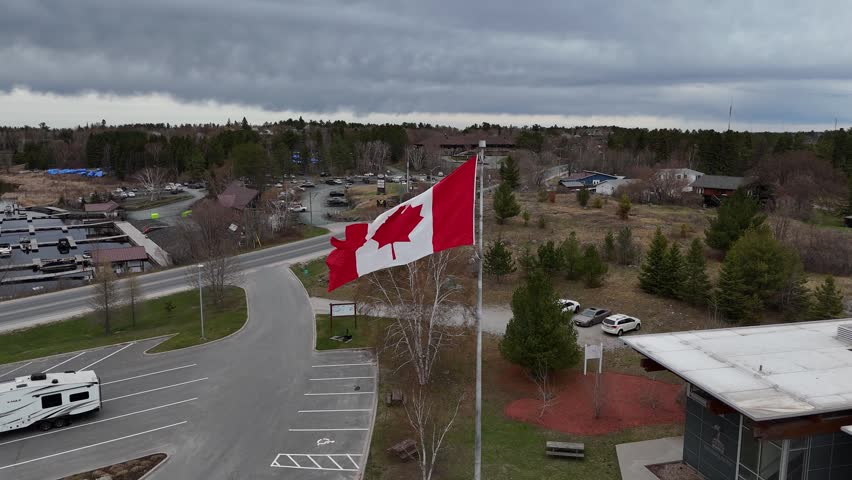 Canadian flag in Kenora, Ontario on Lake of the Woods. Dronie shot backs over water revealing marina, town shoreline, highway along lakeside, boats, and cars on a spring day.