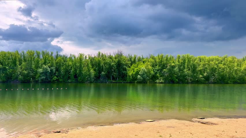 Calm forest lake under dramatic storm clouds. Gloomy dark sky before thunderstorm over green summer woodland. Static wide shot of a peaceful green emerald color lake water with sandy shore and lush forest line. Cinematic ominous overcast clouds before a heavy summer rain.
