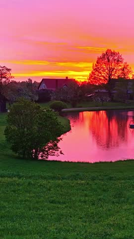 Aerial view moves over pond reflecting vivid sunset beside house and spring trees