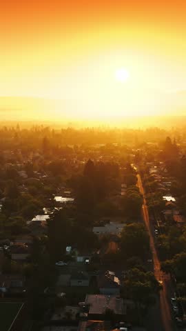 High altitude drone shot gliding over residential houses during glowing sunrise. Vibrant orange sky fills frame while morning mist settles over green trees and suburban streets in slow motion.