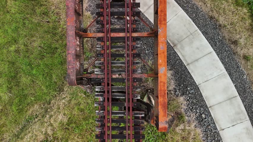 A bike pathway winding below an old rusty rail bridge spanning over a natural flowing creek and pedestrian bridge. Drone view
