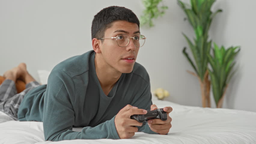 Man lying on bed holding a joystick with clenched fists, smiling and wearing glasses beside a gamepad on a white duvet in a studio; excitement.