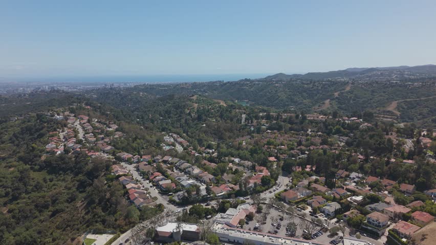 Dense residential neighborhood of red-roofed homes winds along tree-covered hillside canyons with Pacific Ocean and hazy skyline beyond in Beverly Hills, California, USA. Aerial drone wide shot