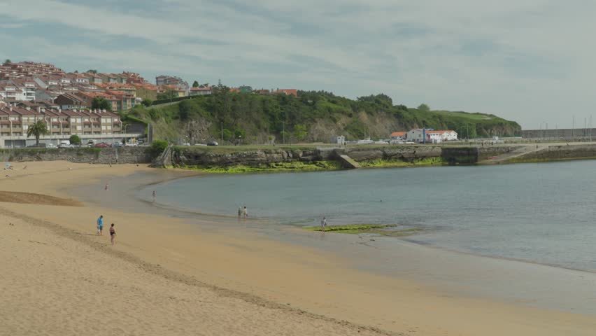 Lovely medium-wide shot of the golden sand beach cove in Luanco, Spain. Features beachgoers along the calm ocean shoreline with traditional coastal apartment blocks sitting on the hills behind.
