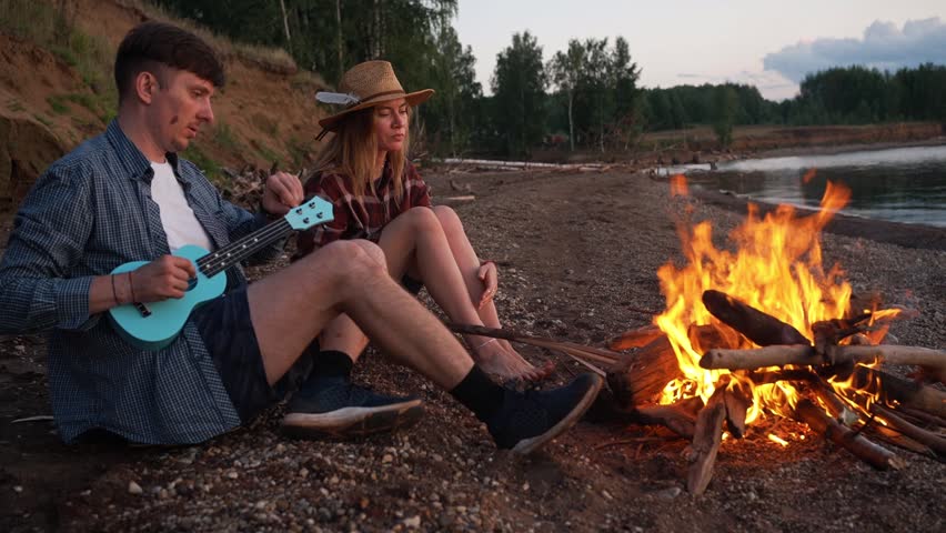 Youthful couple sharing a romantic evening by a river, with the man strumming a ukulele and the woman holding a flickering torch, seated next to a cozy bonfire as the sun sets