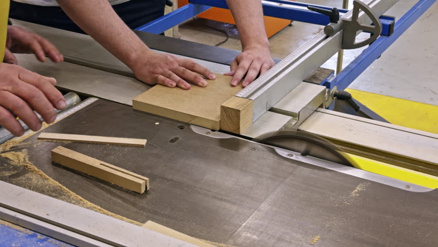 Adult hands are guiding a wooden board through a table saw in a workshop. The clip shows a woodworking task in progress with precision cutting and sawdust visible.