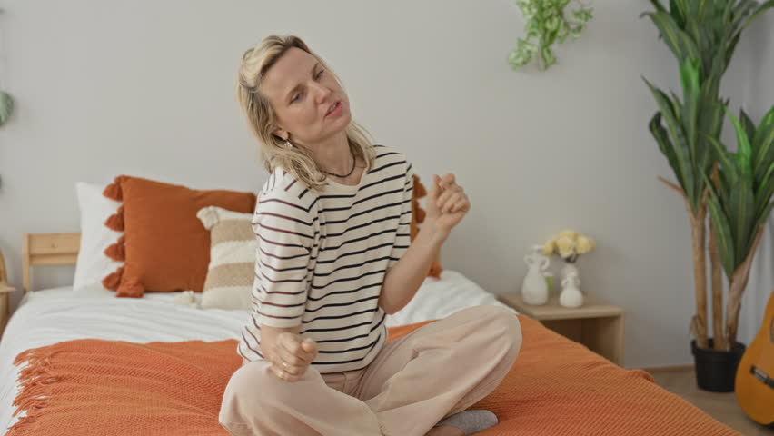 Woman stretching arms on bed, seated crosslegged with raised hands, orange throw and pillows visible in a bright bedroom inside a building; calm contentment.