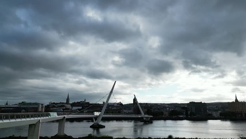 Aerial drone footage of the Peace Bridge spanning the River Foyle in Derry, Northern Ireland, on an overcast evening. The smooth approach highlights the distinctive curved pedestrian bridge, surrounding cityscape and riverside setting under soft evening light.