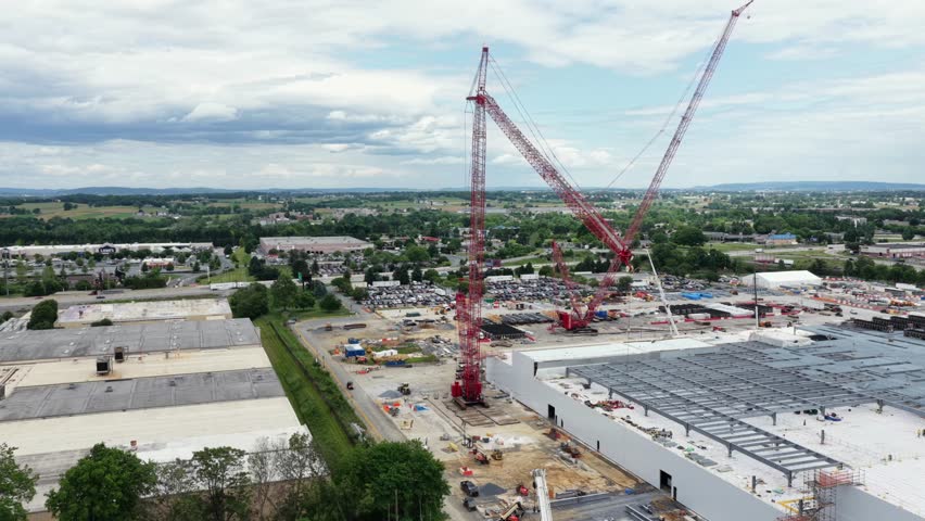 Twin red cranes tower above data center under construction, with steel frame and rolling Pennsylvania farmland under partly cloudy skies in Lancaster, Pennsylvania, USA. Aerial drone wide shot.