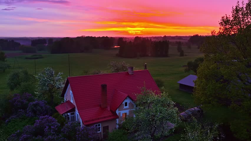 Aerial view of traditional Latvian red roof farmhouse in rural countryside at vivid orange sunset surrounded by green fields blossoming trees and forest with pond
