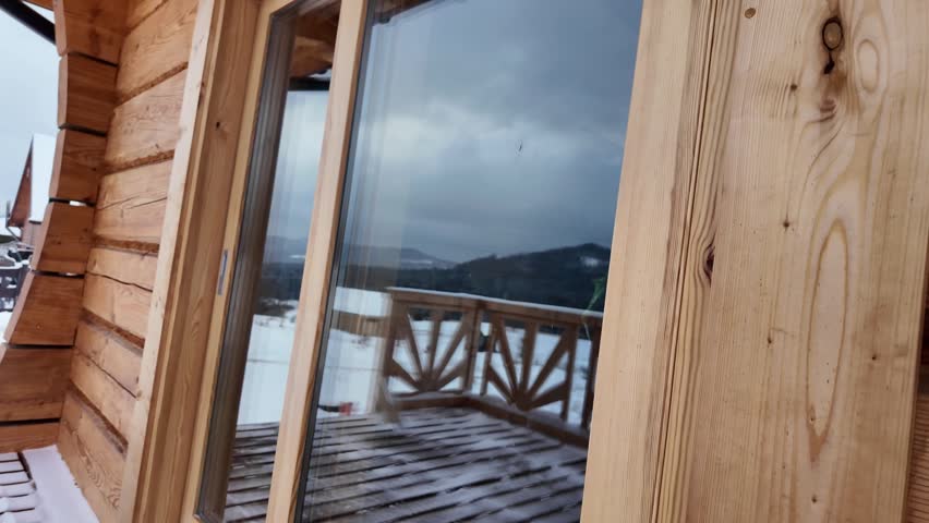 Camera glides along a wooden cabin terrace toward a large window reflecting the railing and snowy mountain peaks