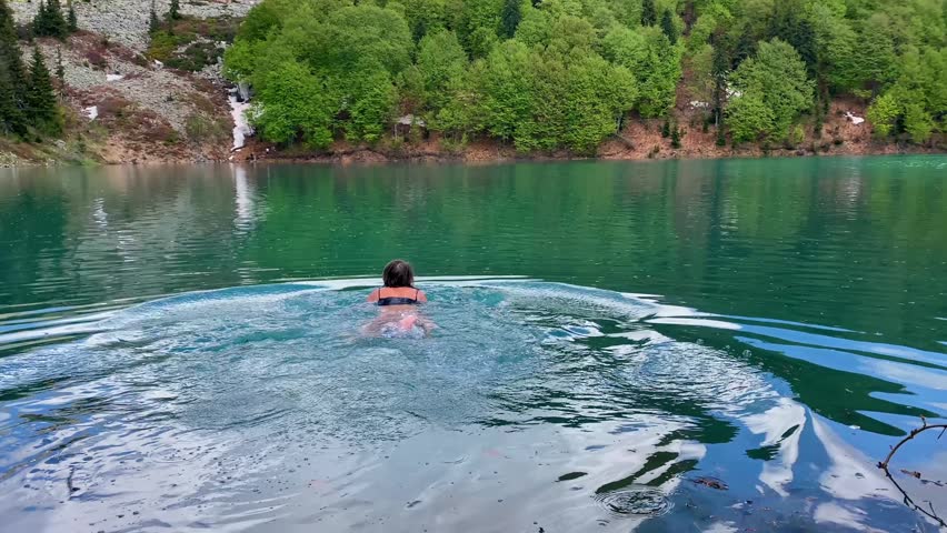 Woman swims across the cold emerald waters of Green Lake in Georgia. Peaceful mountain scenery, forested shoreline, and refreshing outdoor adventure in the Caucasus.