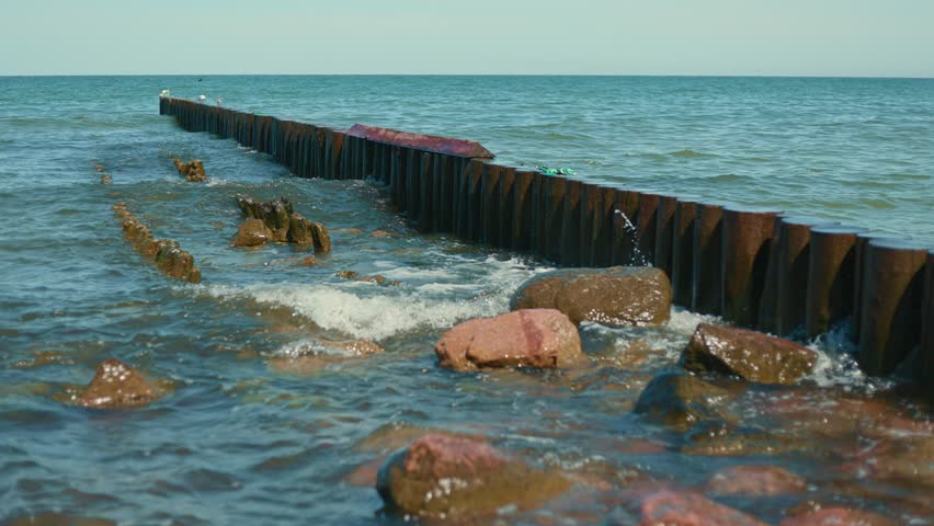Sea waves washing against a wooden breakwater at the beach. Calm water hitting wooden poles and rocks along the shore. Coastal landscape in the daylight time.