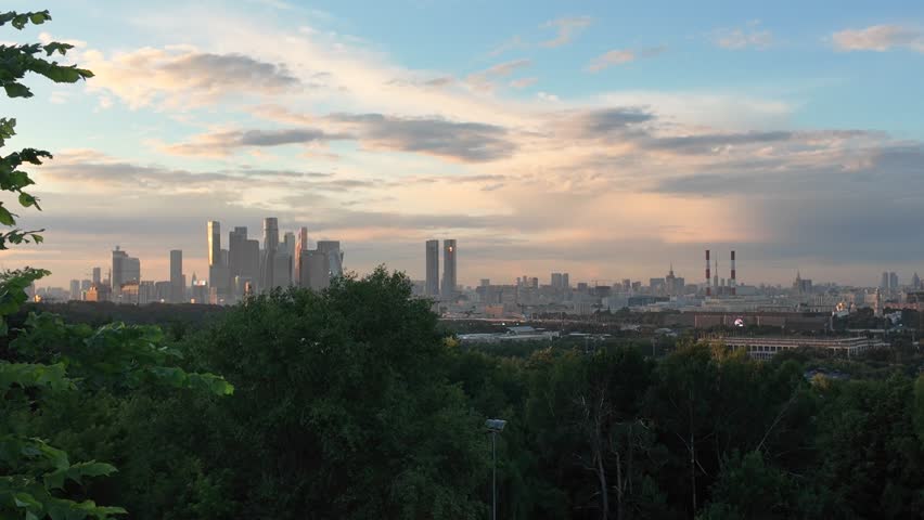 Aerial panoramic view of Moscow city skyline at sunset with dramatic clouds and warm golden light. Modern skyscrapers and urban districts surrounded by green forested areas, showcasing the contrast between nature and one of Europe