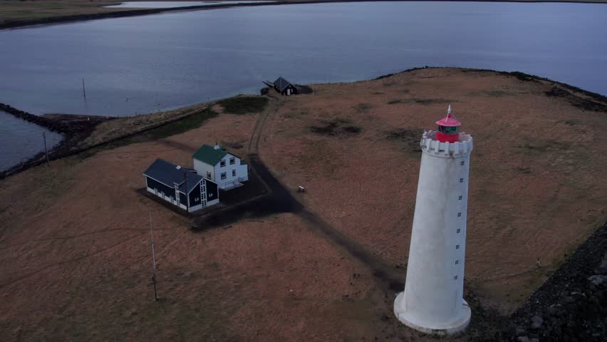 Drone flying over the scenic Grotta lighthouse, a beacon on a peninsula near Reykjavik. The coastal landscape shows traditional Icelandic houses