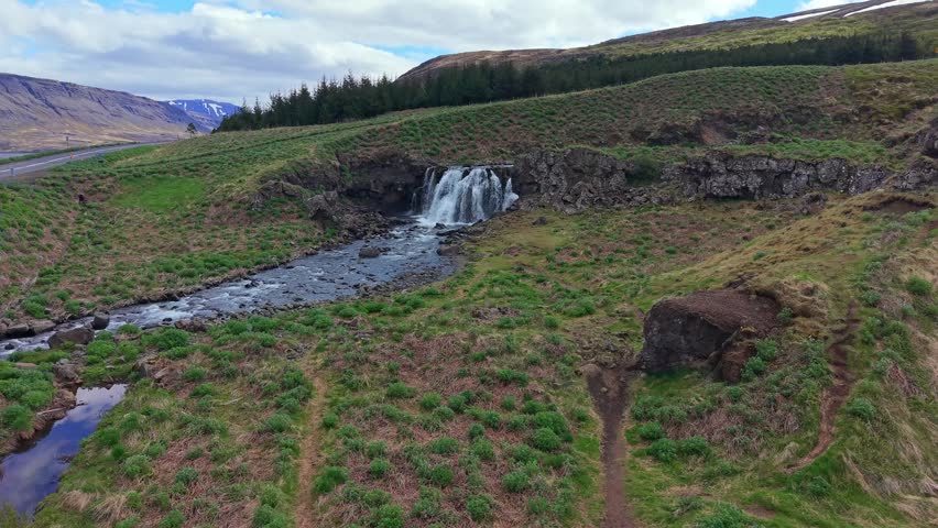 Aerial view approaching the majestic Fossarett waterfall, a beautiful cascade flowing through the lush green landscape of Iceland