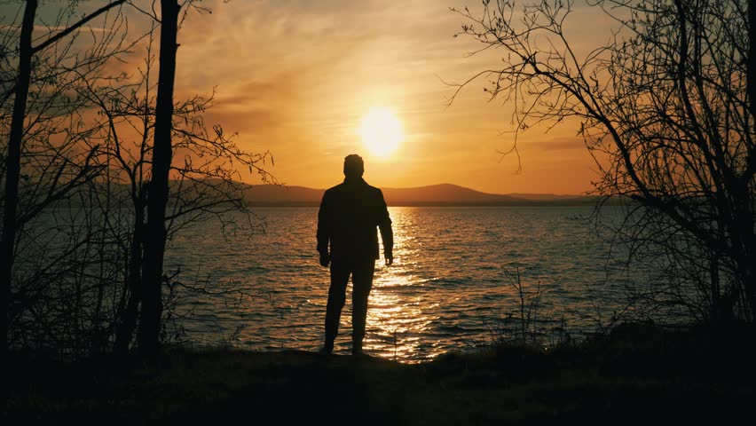 The silhouette of a lonely man who stands quietly and admires the sunset on the shore of the lake. Harmony of nature and man, time alone with oneself.	