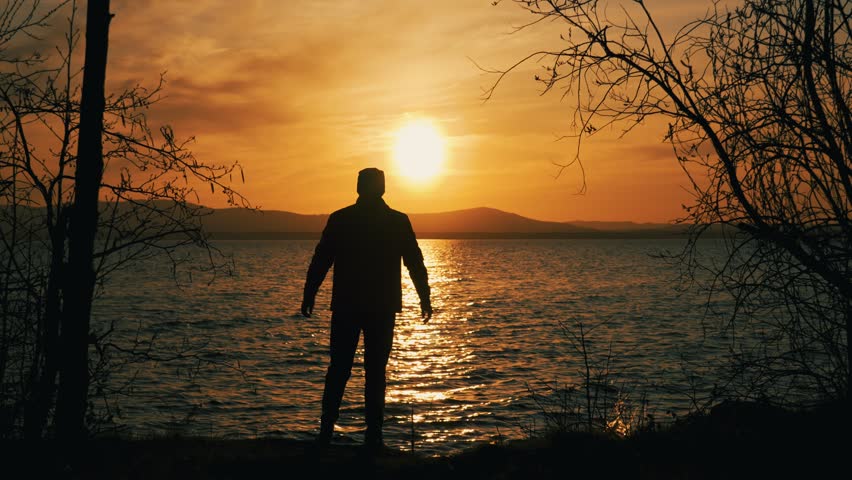 The silhouette of a single man standing on the shore of a lake at sunset. A happy person enjoys life, achievements and success by raising his hands in the air. Rear view.	