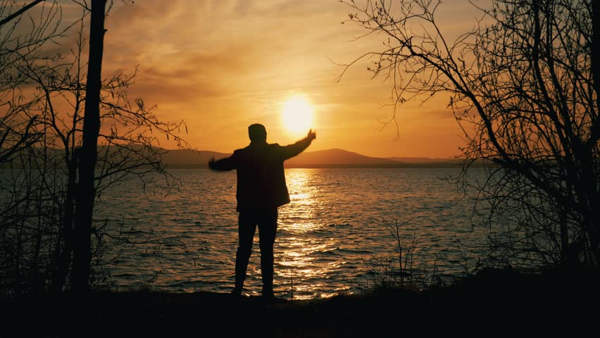 Silhouette of a man standing on the shore of a lake at sunset. He waves his hands, indicating his place or asking for help.